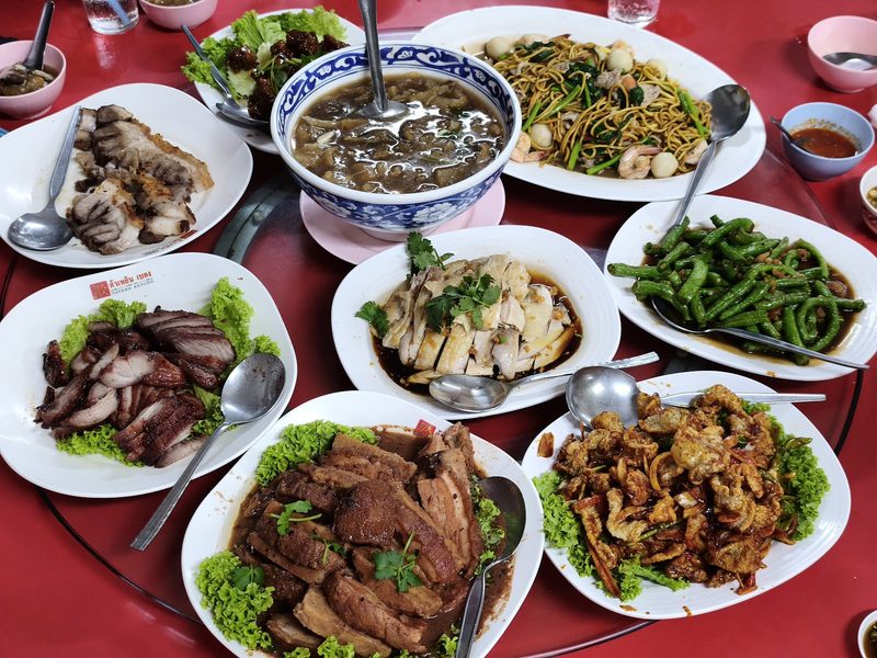 อาหารจีนหลายจานเต็มโต๊ะกลมผ้าแดง มีหมูแดง ไก่สับ ถั่วเจี๋ยน และหมี่ผัด